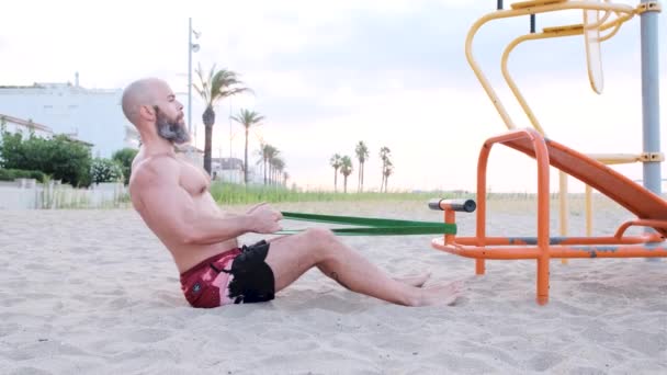 Uomo sportivo che fa allenamento di calistenica con un elastico di resistenza mentre seduto sulla sabbia in spiaggia. Sport e fitness concept. — Video Stock