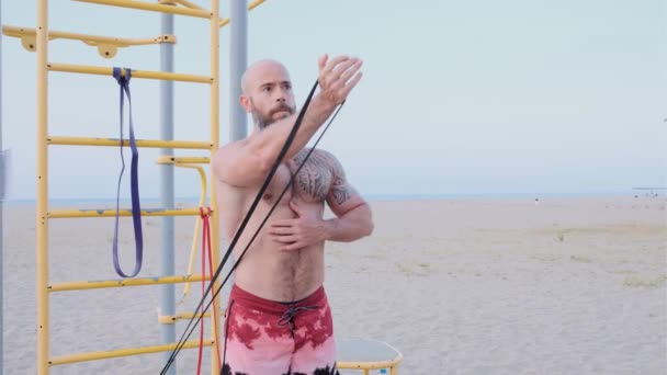 Fitness man gör biceps triceps calisthenics träning med ett motstånd gummiband på stranden. Idrottsbegreppet. — Stockvideo