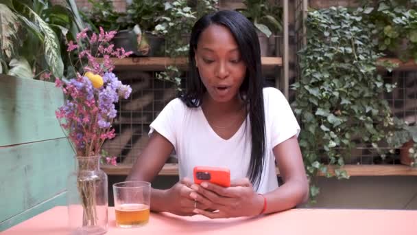 Young woman surprised by news and using a mobile phone and smiling while sitting in a restaurant or coffee shop garden. — Stock Video