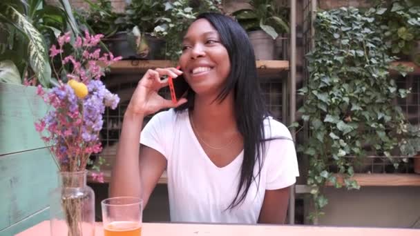 Young woman using a mobile phone and laughing while sitting in a restaurant or coffee shop garden. — Stock Video