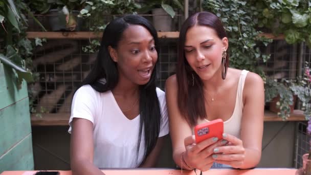 두 명의 젊은 친구가 커피 샵이나 식당 정원에 함께 앉아 스마트폰을 사용하고 웃고 있습니다. 우정 개념. — 비디오