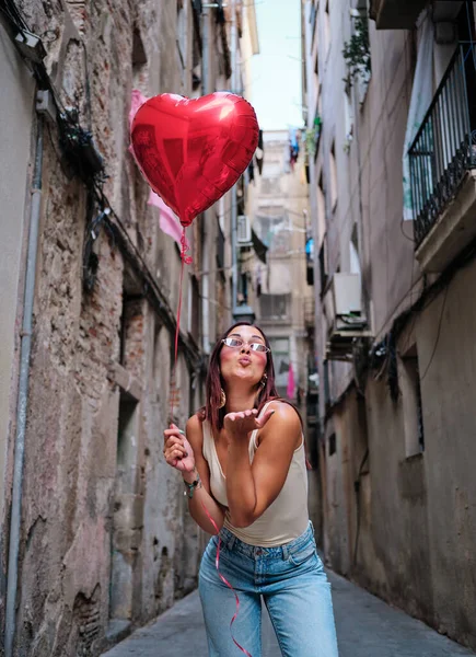 Donna che soffia un bacio d'aria alla fotocamera mentre tiene un palloncino rosso a forma di cuore all'aperto. — Foto Stock