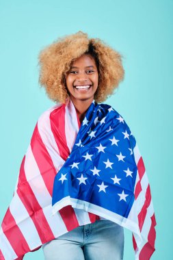 İzole edilmiş bir arka planda ABD bayrağıyla sarılmış gülümseyen genç bir afro kadın..