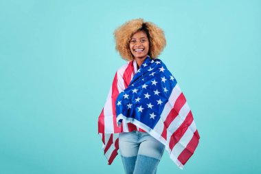 İzole bir arka planda dururken Amerikan bayrağına sarılmış gülümseyen afro kadın..