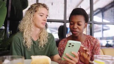 Modern bir restoranda yemek yerken cep telefonu ekranına bakıp gülümseyen ve birlikte gülen çok ırklı kadın arkadaşlar.