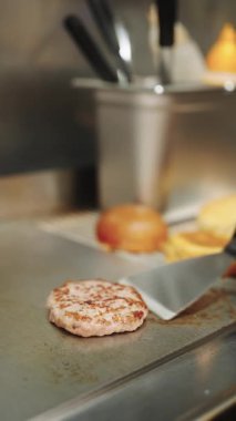 Siyah eldivenli profesyonel bir aşçı ticari bir mutfak ızgarasında sulu bir köfteyi çeviriyor. Arka planda çörekler kızartılıyor.