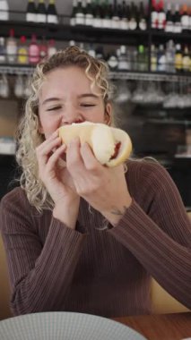 Lokantada oturan neşeli sarışın kadın patates kızartmalı sosisli sandviçi mutlulukla yiyor. Fast food konsepti ve dışarıda yemek yemek.