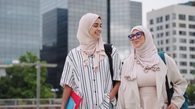 İki genç Müslüman kadın başörtüsü takıyor ve arka planda modern binalar olan bir üniversite kampüsünde mutlu mesut yürüyüp konuşuyorlar.