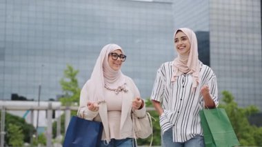 İki neşeli genç Müslüman kadın alışveriş torbalarıyla birlikte yürüyorlar. Modern bir şehirde muhabbet eden ve gülen mutlu bayan arkadaşlar.