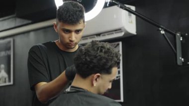 Genç profesyonel berber, kıvırcık saçlı müşterilerine tıraş bıçağı kullanıyor. Şık bir berber dükkanında modern bir saç stili.