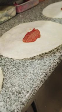 Profesyonel bir pizzacının, siyah eldiven giyip çiğ bir pizza tabanına taze domates sosu sürerken dairesel bir hareketle kaşıkla yakın plan fotoğrafı.