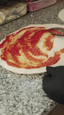 Siyah eldivenli profesyonel bir şef çiğ pizza hamuruna kaşıkla taze domates sosu sürüyor. Restoran mutfağında pizza pişirme işlemi