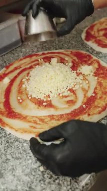 Siyah eldivenler giyen erkek pizzacı, domates sosunu dikkatlice yayıyor ve pizzacıda çiğ pizza hamuruna mozzarella peyniri ekliyor.