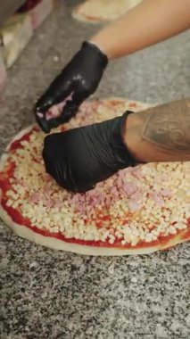 Profesyonel şeflerin elleri siyah eldivenlerle mozzarella ve domates sosunun üzerine doğranmış jambon yayıyor. Çiğ pizza hamuru üzerinde çalışıyorlar.