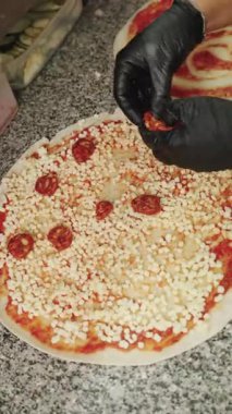 Siyah eldiven giyen profesyonel pizzacı, domates soslu pizzaya pepperoni dilimleri ve taş tezgahta mozzarella peyniri ekliyor.