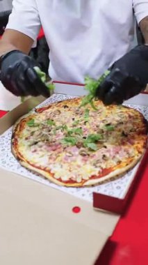 Siyah eldivenler takmış bir pizzacı şefinin fotoğrafını çek. Taze roka yapraklarını paket servis kutusunun içindeki sıcak pizzanın üstüne dikkatlice yerleştir.