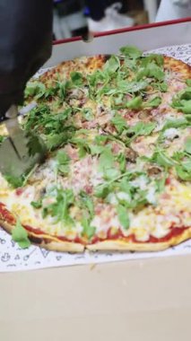 Siyah eldivenli profesyonel aşçı jambonlu, mantarlı ve roka kaplı taze bir pizzayı karton kutuda dilimlemek için patenci kullanıyor.