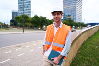 Genç erkek mühendis ya da inşaat işçisi güvenlik yeleği ve başlık takıyor, kentsel bir ortamda dijital tablet tutuyor.