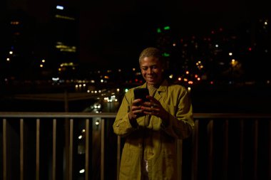 Bir kadın gülümsüyor, akıllı telefonuyla sosyal medyayı kontrol ediyor, şehir ışıkları ve bokeh ile bir balkonda duruyor.