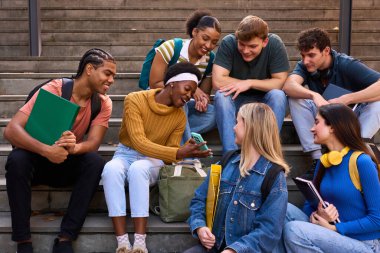 Çeşitli öğrenci grupları üniversite kampüsünün merdivenlerinde otururken sosyalleşiyorlar, konuşuyorlar ve birlikte akıllı telefona bakıyorlar.