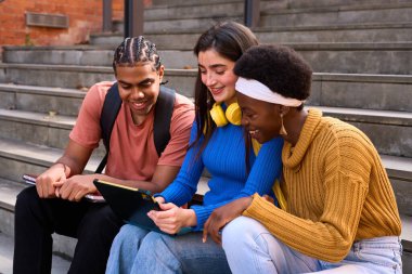 Dijital tablette birlikte çalışan, fikirleri paylaşan ve kampüs merdivenlerinde gülümseyen bir grup mutlu genç öğrenci.