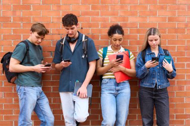 Bir grup genç öğrenci, turuncu bir duvarın önünde dururken birbirlerini görmezden gelerek akıllı telefonlarına odaklandılar.