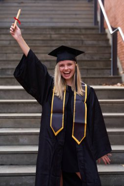 Üniversite mezuniyetini kutlayan genç bir kız öğrenci, elinde diploma tomarı, merdivenlerde duruyor. Neşeli sarışın kadın akademik başarı elde ediyor
