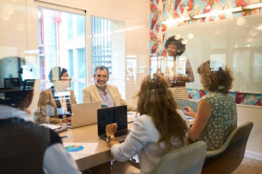 Modern bir ofis yönetim kurulundaki toplantı sırasında çeşitli iş adamları bir sunucuyu dinleyip işbirliği yapıyor.