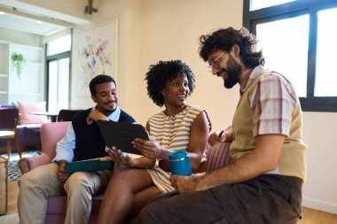 Gayri resmi ofis ortamında oturan çeşitli iş arkadaşları, dijital tablet üzerinde çalışırken gülümseyip iş konuşuyorlar.