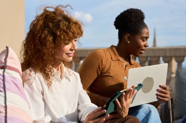 İki farklı iş kadını, güneşli bir teras ofisinde dizüstü bilgisayar ve tablet üzerinde birlikte çalışıyor.