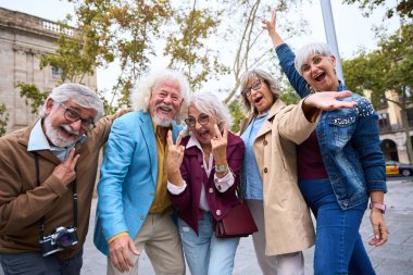 Bir grup mutlu kıdemli arkadaş, eğlenceli tatillerinde şehirde selfie çekerken gülümsüyor ve barış işareti yapıyor.