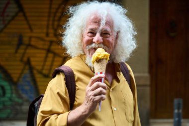 Mutlu son sınıf öğrencisi, beyaz saçlı, neşeli bir şekilde gülümseyen ve şehir caddesinde leziz bir dondurma külahı yiyen.