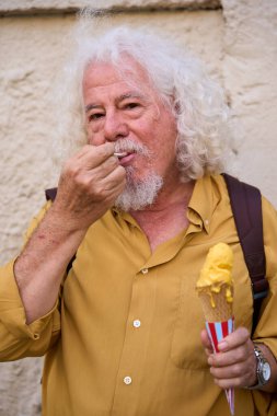 Beyaz sakallı ve sırt çantalı yaşlı adam güneşli bir sokakta sarı dondurma külahının tadını çıkarıyor. Rahatlamış, mutlu bir yaz portresi, samimi bir seyahat anı.