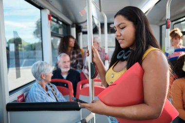 Genç bir kız öğrenci elinde bir dosya tutuyor ve her gün otobüste dururken akıllı telefonunu kullanıyor.