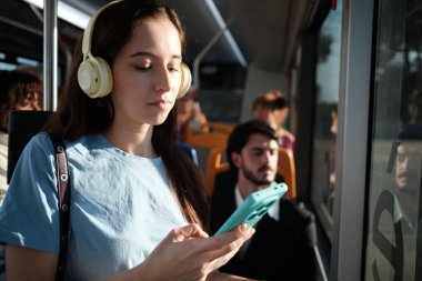 Genç bir kadın toplu taşıma aracına biniyor, ses içeriğini dinliyor ve cep telefonunu kullanıyor. Her gün seyahat eden yolcu