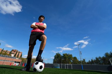 Kendine güvenen genç futbolcu yeşil bir yapay çim sahasında duruyor mavi gökyüzünün altında bir ayağı topun üzerinde.