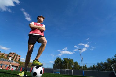 Kendine güvenen genç bir sporcu parlak mavi gökyüzünün altında futbol sahasında poz veriyor, antrenmana hazırlanıyor.