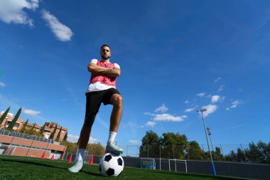 Kolları çapraz duran, futbol topuna basan kendine güvenen futbolcu, mavi gökyüzünün altındaki yapay çim sahasında.