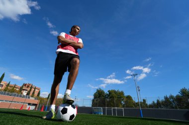 Kendine güvenen genç bir erkek atlet, açık mavi gökyüzünün altındaki suni çim sahasında bir futbol topunun üzerinde duruyor.