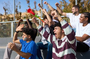 Çeşitli gençler coşkuyla tezahürat yapıyor, yumruklarını kaldırıyor ve bir spor müsabakası sırasında vuvuzela üflüyorlar.