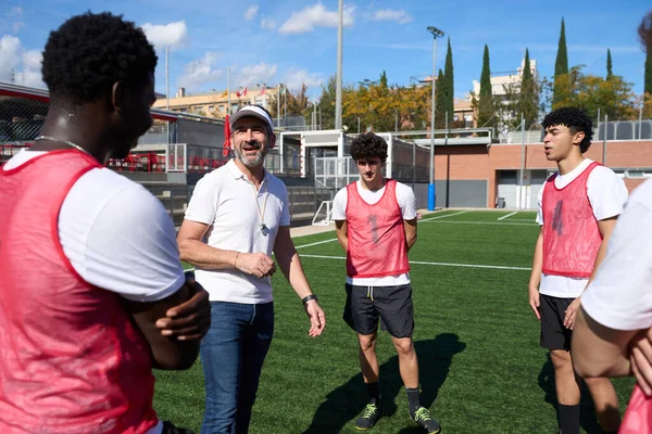 Futbol koçu, güneşli bir futbol sahasındaki antrenman sırasında çeşitli genç erkek oyunculara talimat veriyor.