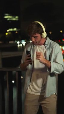 Şehir balkonunda dans ederken kulaklık takan ve elinde akıllı telefon tutan neşeli genç bir adam. Bulanık bir şehir gece hayatı arka planına karşı ritme ayak uyduruyor.