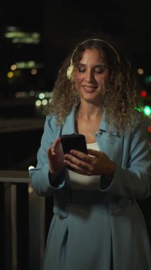 Şık, kıvırcık saçlı, kulaklıklarıyla müzik dinleyen genç bir kadın. Akıllı telefonunu kullanırken gülümsüyor ve geceleri şehir balkonunda dans ediyor.