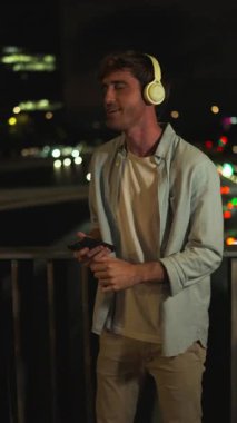 Kulaklık takan neşeli genç adam, akıllı telefonundan müzik dinliyor ve Bokeh ışıkları olan bir köprüde mutlu bir şekilde dans ediyor.