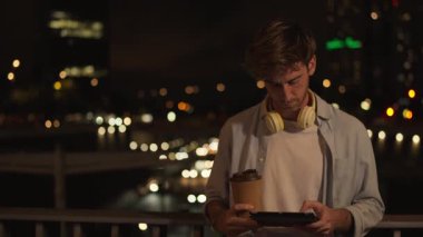 Kulaklıklı sıradan bir genç adam dijital tablet kullanıyor ve gecenin bir yarısı Bokeh City ışıklarıyla şehir balkonunda kahve fincanı tutuyor.