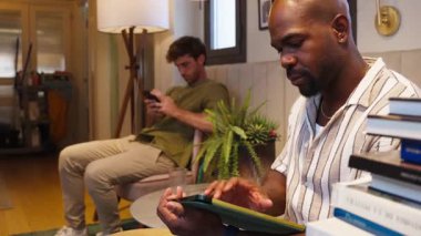 Oturma odasında elektronik aletlerini kullanan iki çok ırklı adam. Siyah bir adam tablet kullanıyor ve arkadaşı mesaj atıyor.