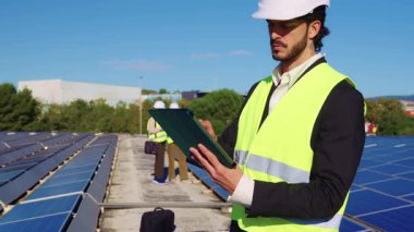 Çatı katındaki güneş enerjisi santralinin bakım denetimi için dijital tablet kullanan çelik yelek ve başlıklı odaklanmış erkek mühendis.
