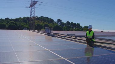 Genç teknisyen sıkı bir şapka ve güvenlik yeleği giyiyor. Yakınlarda elektrik direği olan bir çatı katı güneş enerjisi santralinde bakım yapıyor.