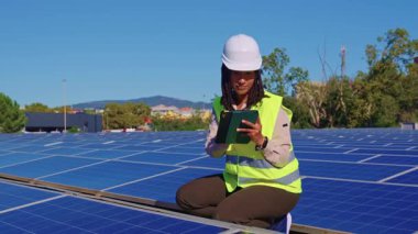 Afrika kökenli Amerikalı kadın mühendis çatıda çalışıyor, fotovoltaik hücreleri güneş enerjisi istasyonunun bakımı için tabletle kontrol ediyor.