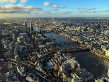 Londons kentsel manzarası ufka kadar uzanıyor, açık bir gökyüzü altında Thames nehri, köprüler ve ikonik binalar yer alıyor.